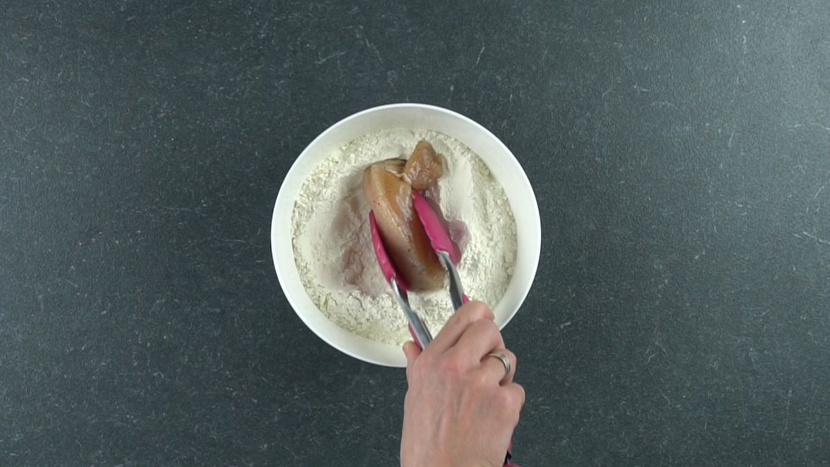 pink tongs dipping chicken into seasoned flour