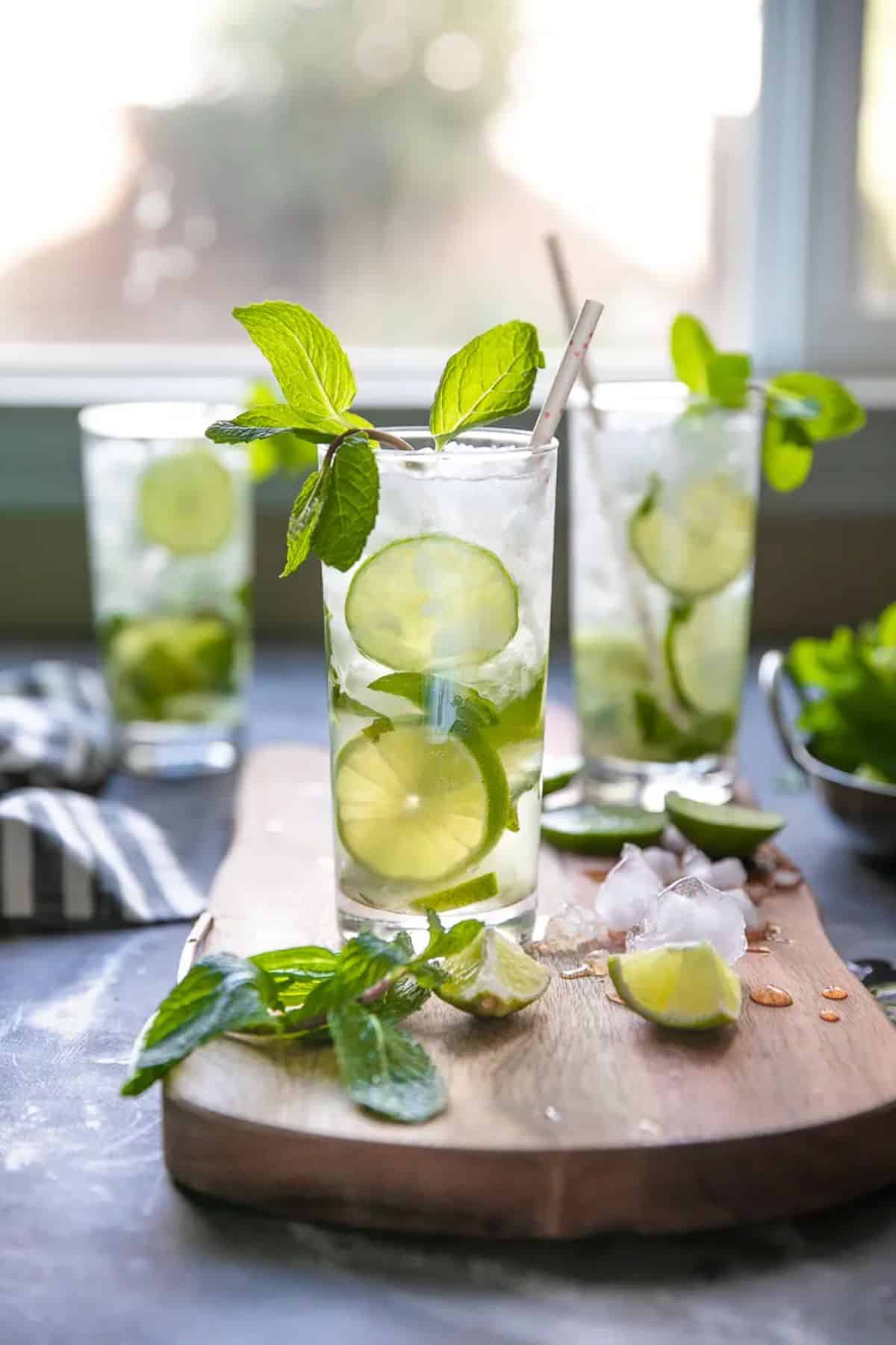 Mojito rum cocktails in two tall glass cups with sliced of lime on a wooden cutting board.
