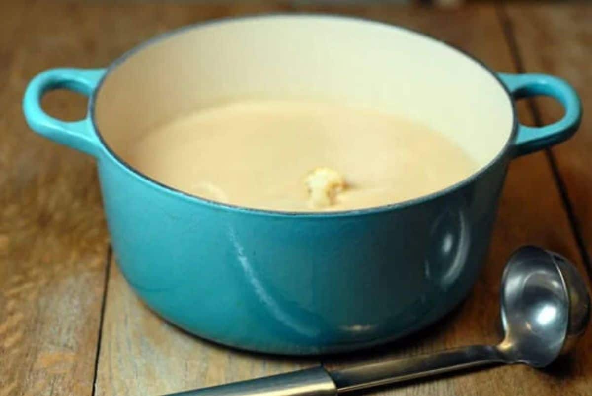 Vegan Roasted Cauliflower Soup in a blue pot on a wooden table with a ladle.