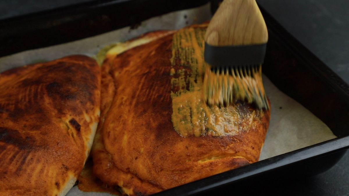 pastry brush adding sauce to top of calzones on baking sheet