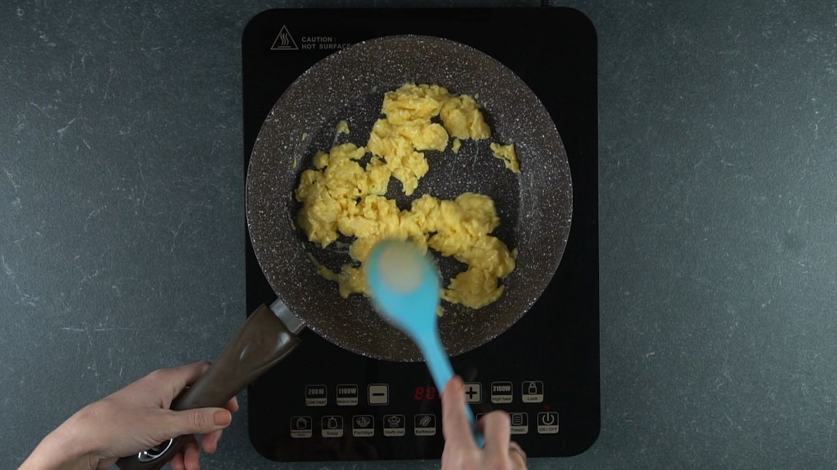 blue spoon stirring cooked eggs in skillet on hot plate