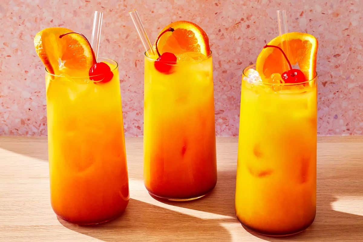Three Tequila Sunrise coctails in tall glasses with straws, slices of orange and cherries.