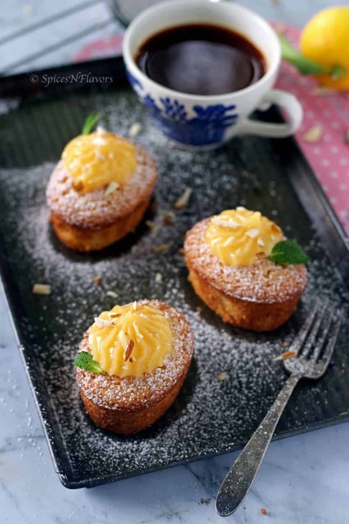Lemon Friands with Lemon Curds on a black tray with a fork and a cup of coffee.