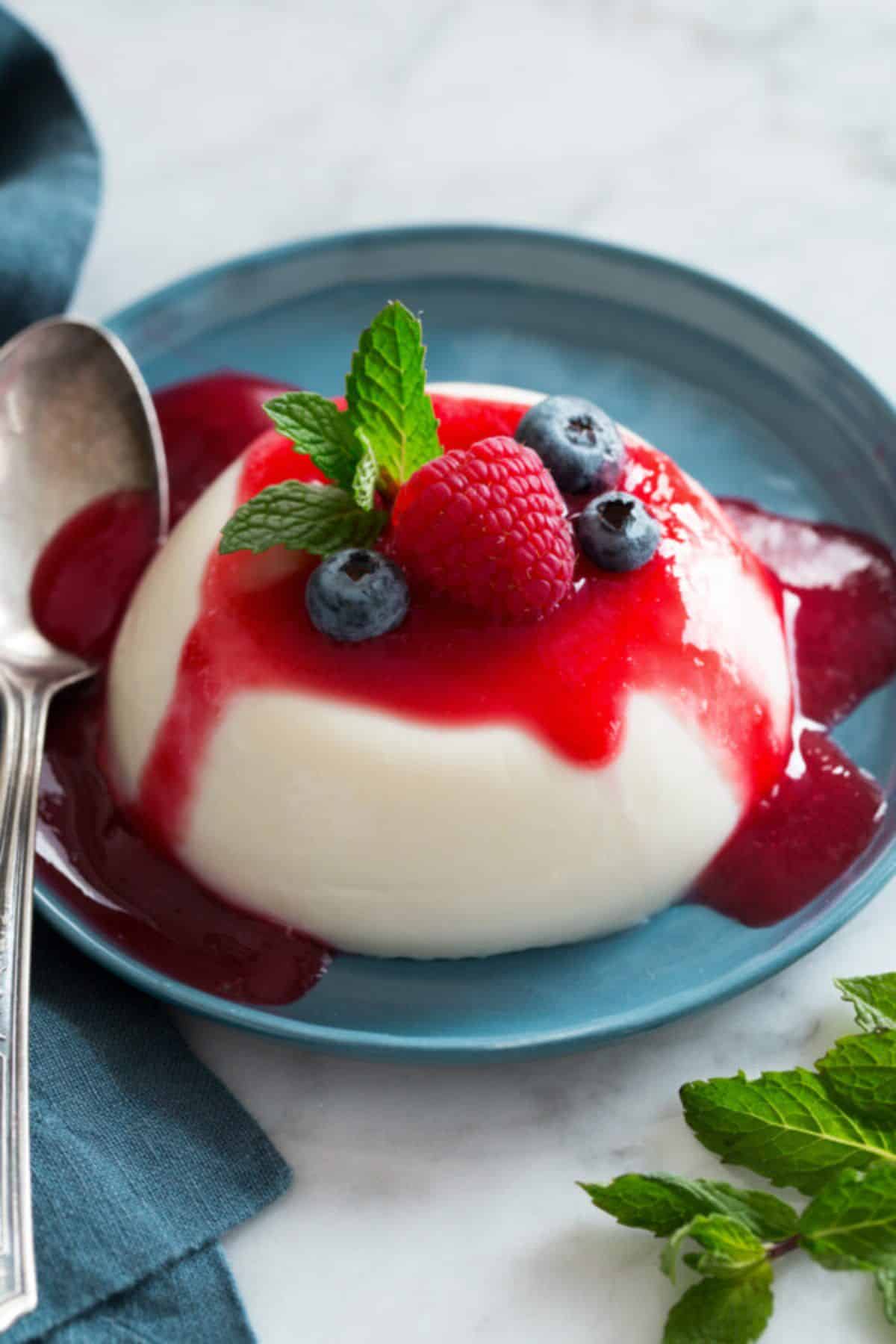 Panna Cotta italian dessert with a strawberry and blueberries on the top on a blue plate with a spoon.
