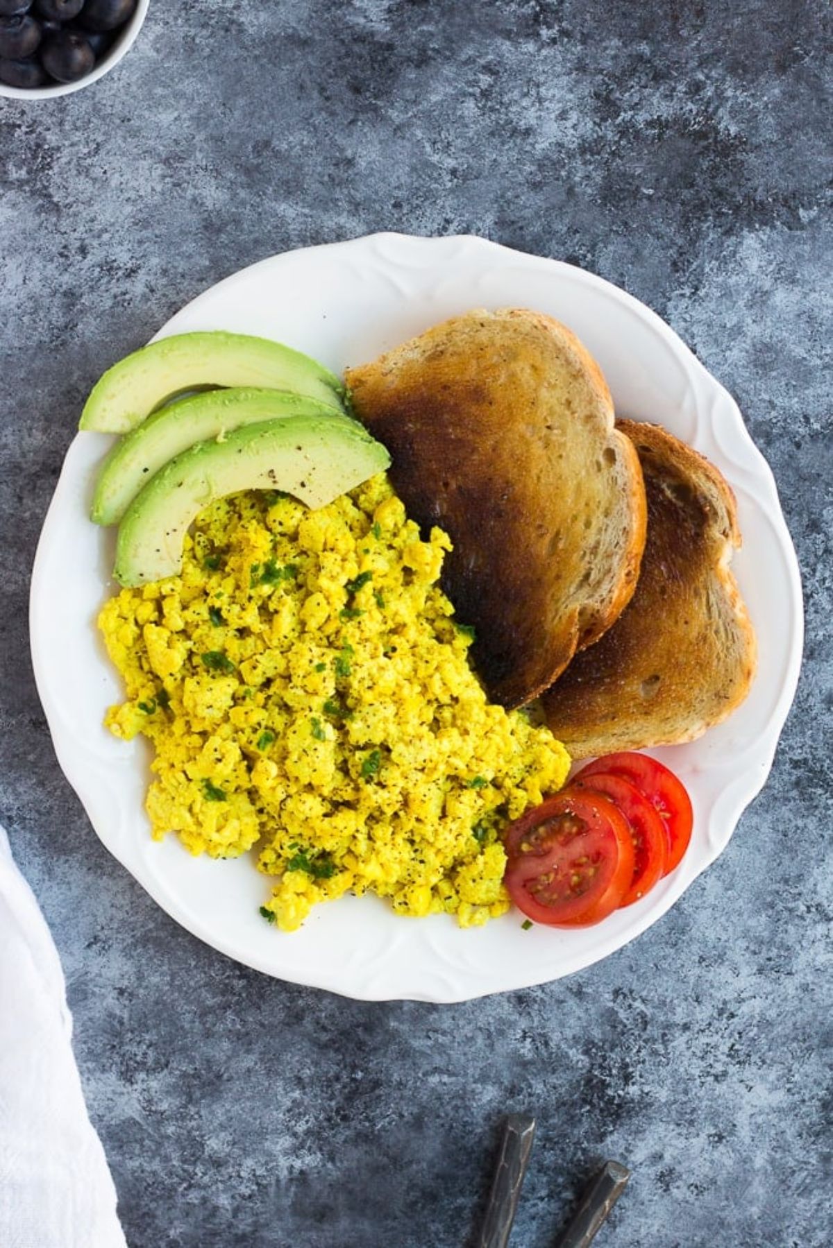 Tofu Scramble with a sliced avocado, tomato and two pieces of bread on a white plate.