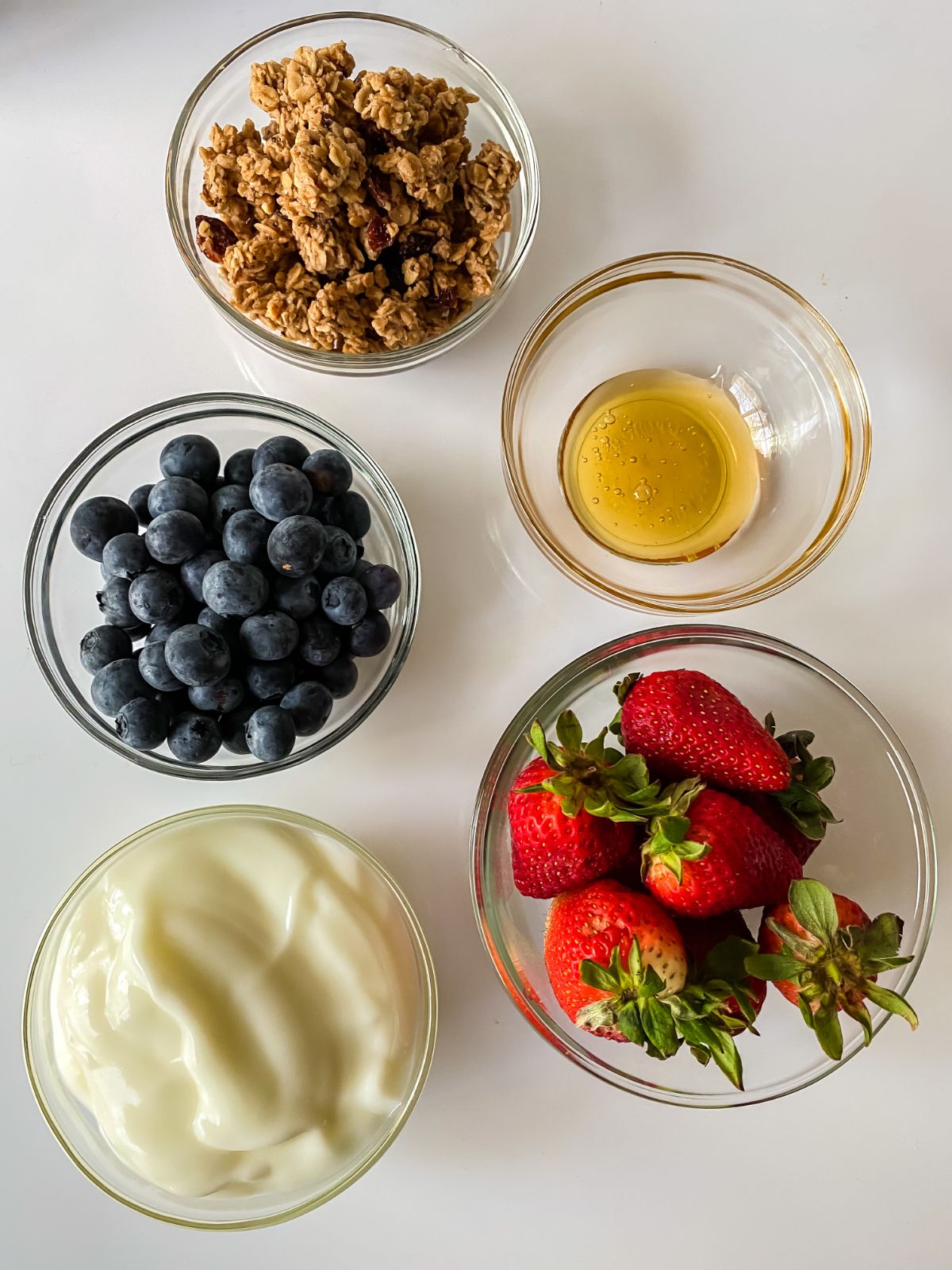 glass bowls of fruit, honey, granola, and yogurt on white table