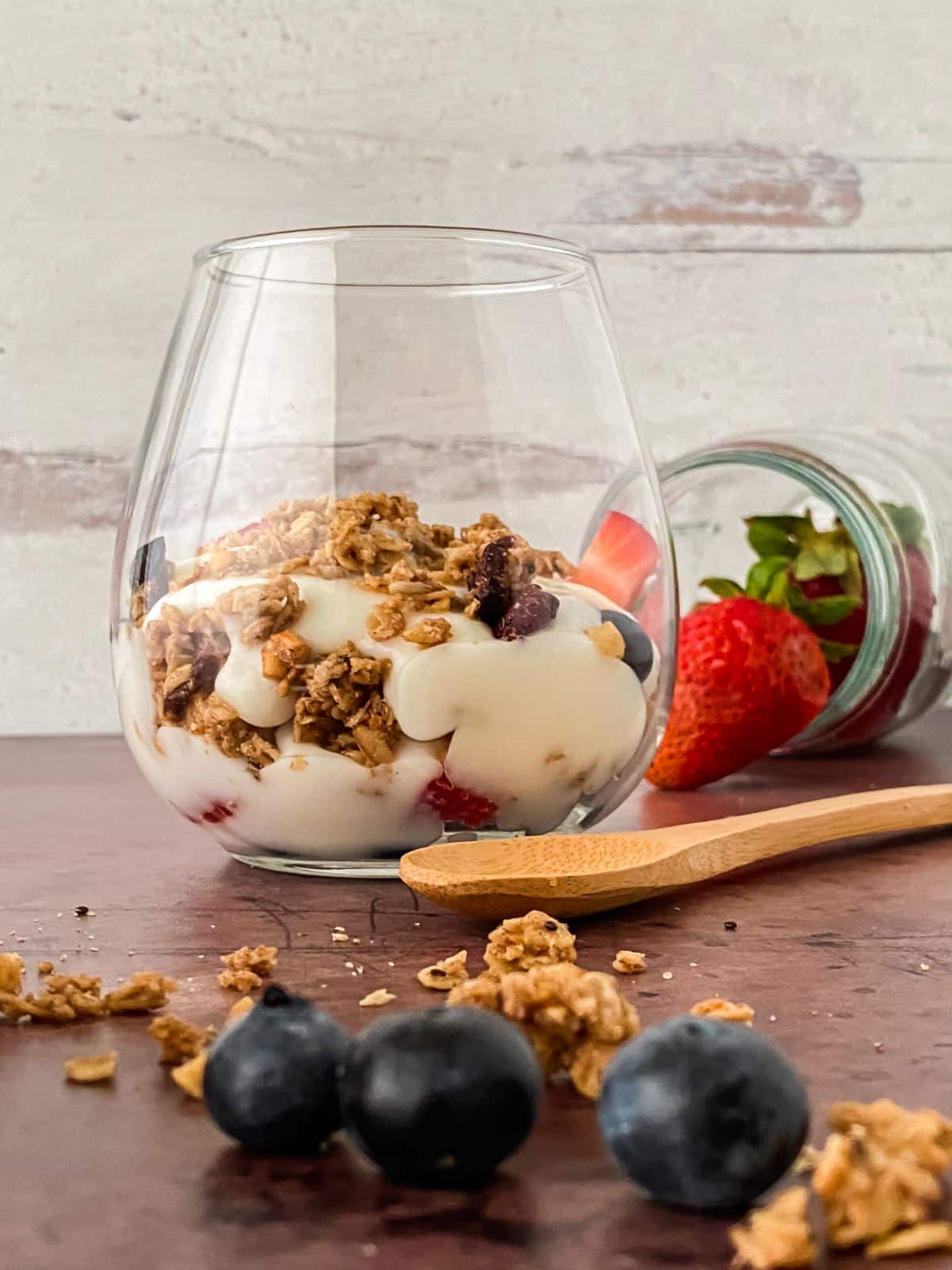 yogurt layered with berries and granola in short glass on table by white wood wall